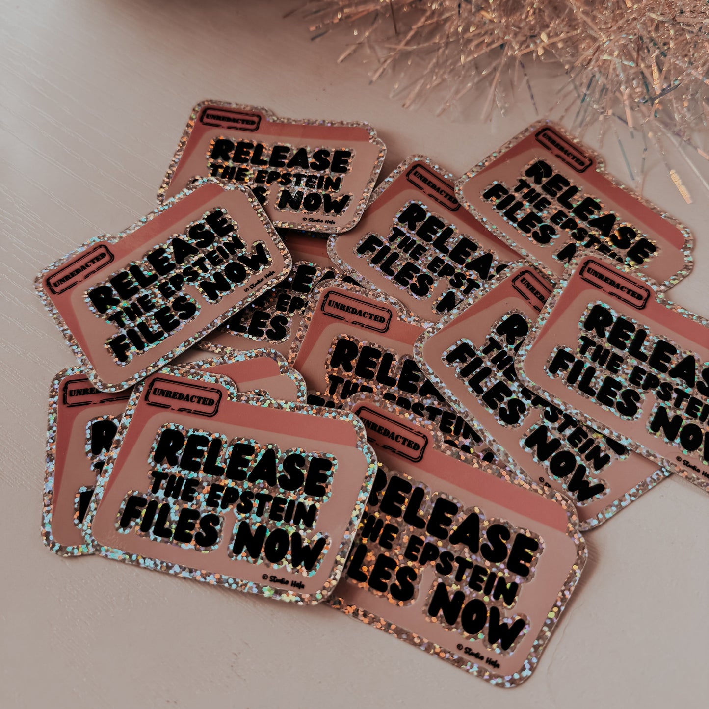 Stickers with 'Release the Epstein files now' text on a reflective surface with decorative elements.
