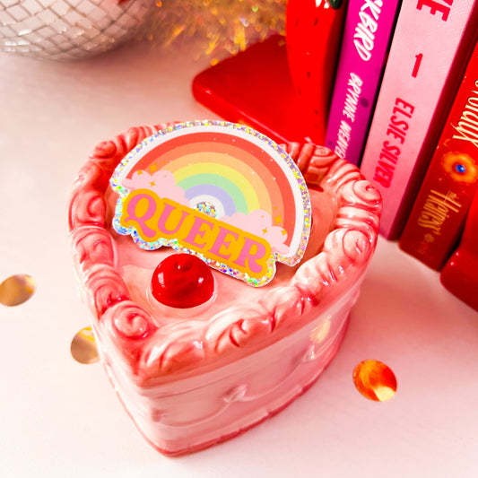 This image displays a set of "Queer/Ally" themed 3-inch glitter vinyl stickers, available in various designs. The image shows two stickers placed on a table among scattered confetti, with one sticker featuring a pink and orange rainbow design, and the other displaying the word " Queer " in bold, black letters against a rainbow background.