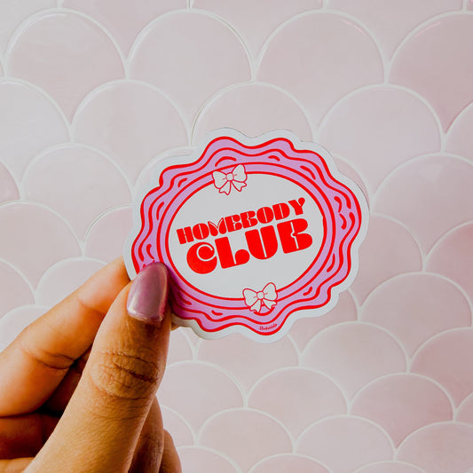 Hand holding a 'Homebody Club' sticker against a patterned background