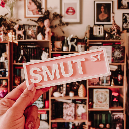 Hand holding a 'SMUT ST' sign in a room with bookshelves and decorative items.