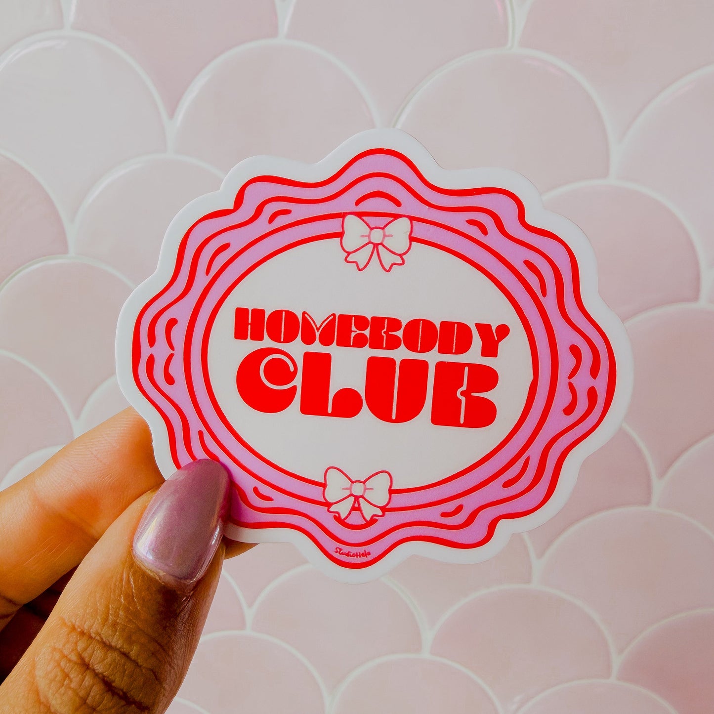 Sticker with 'Homebody Club' text held by a hand against a patterned background