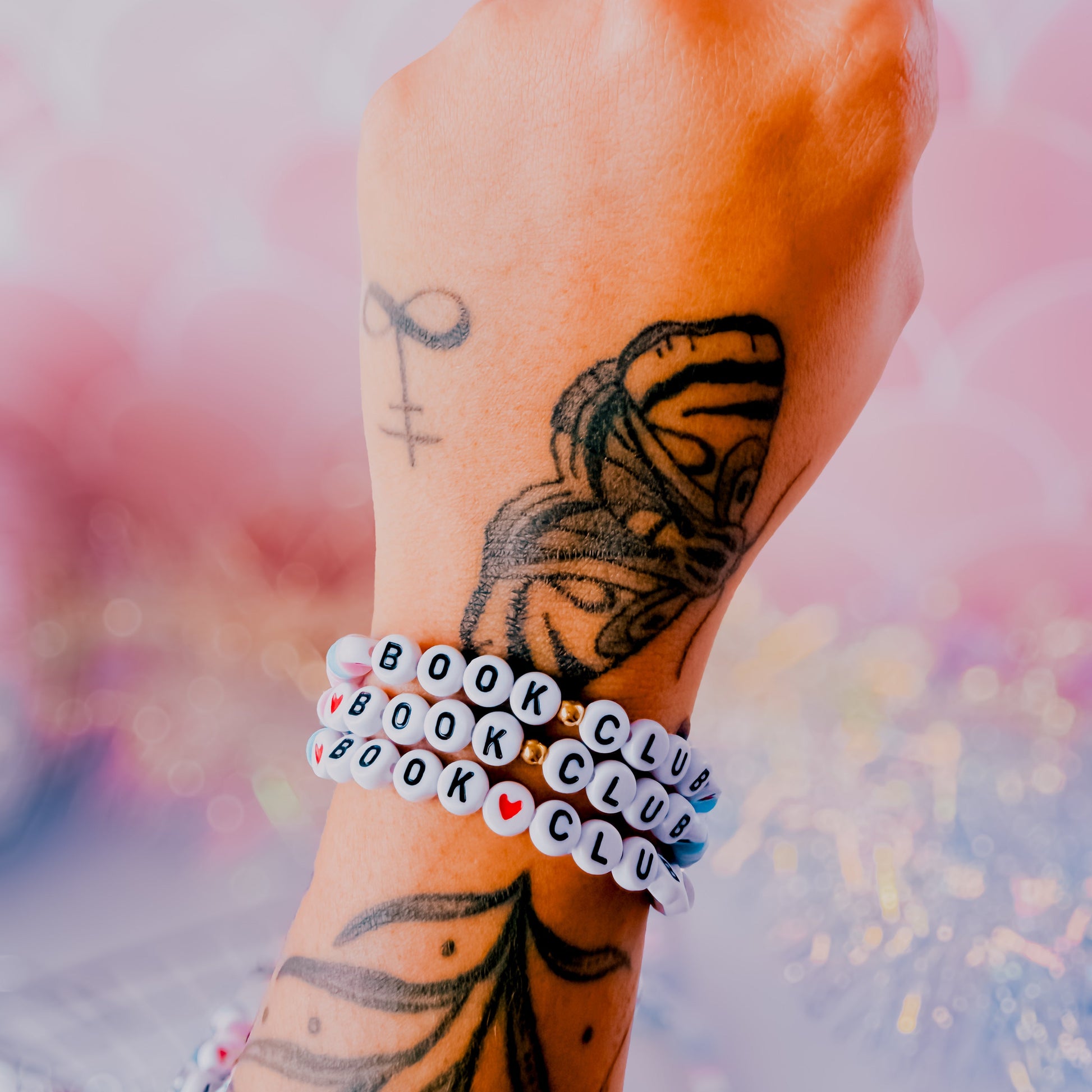 Hand with butterfly tattoo wearing a 'Book Club' bracelet against a blurred background