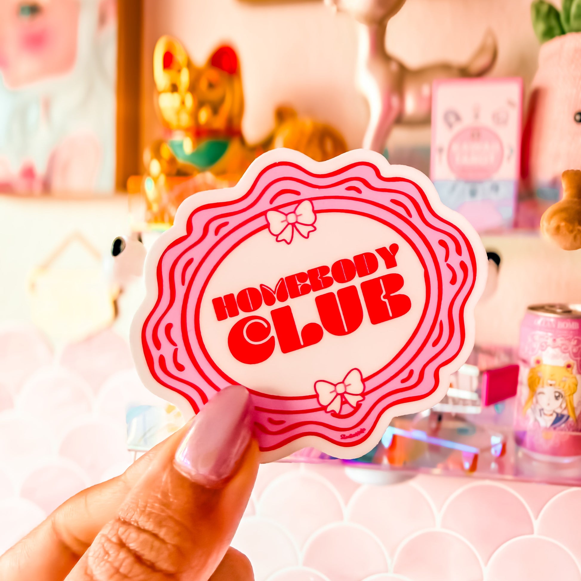 Hand holding a 'Homebody Club' sticker with a decorative background featuring plush toys and stationery.