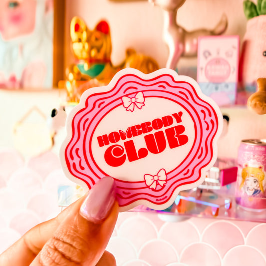 Hand holding a 'Homebody Club' sticker with a decorative background featuring plush toys and stationery.