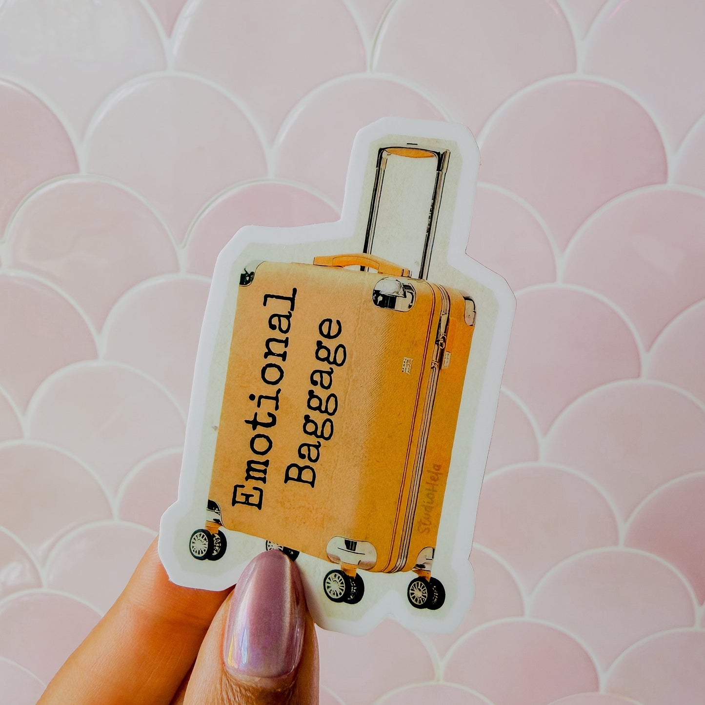 Sticker of a suitcase labeled 'Emotional Baggage' held by a hand against a pink patterned background.