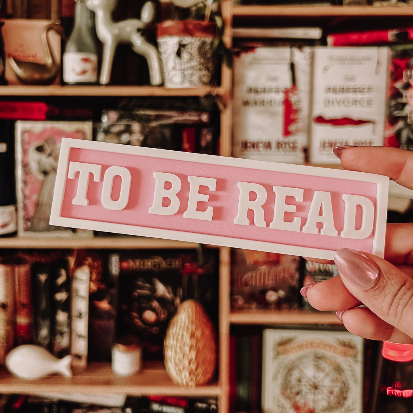 Hand holding a 'TO BE READ' sign in front of a bookshelf filled with books and decorative items.