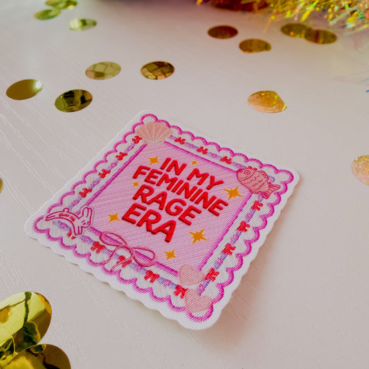Embroidered patch with 'In My Feminine Rage Era' text on a white background with gold confetti.