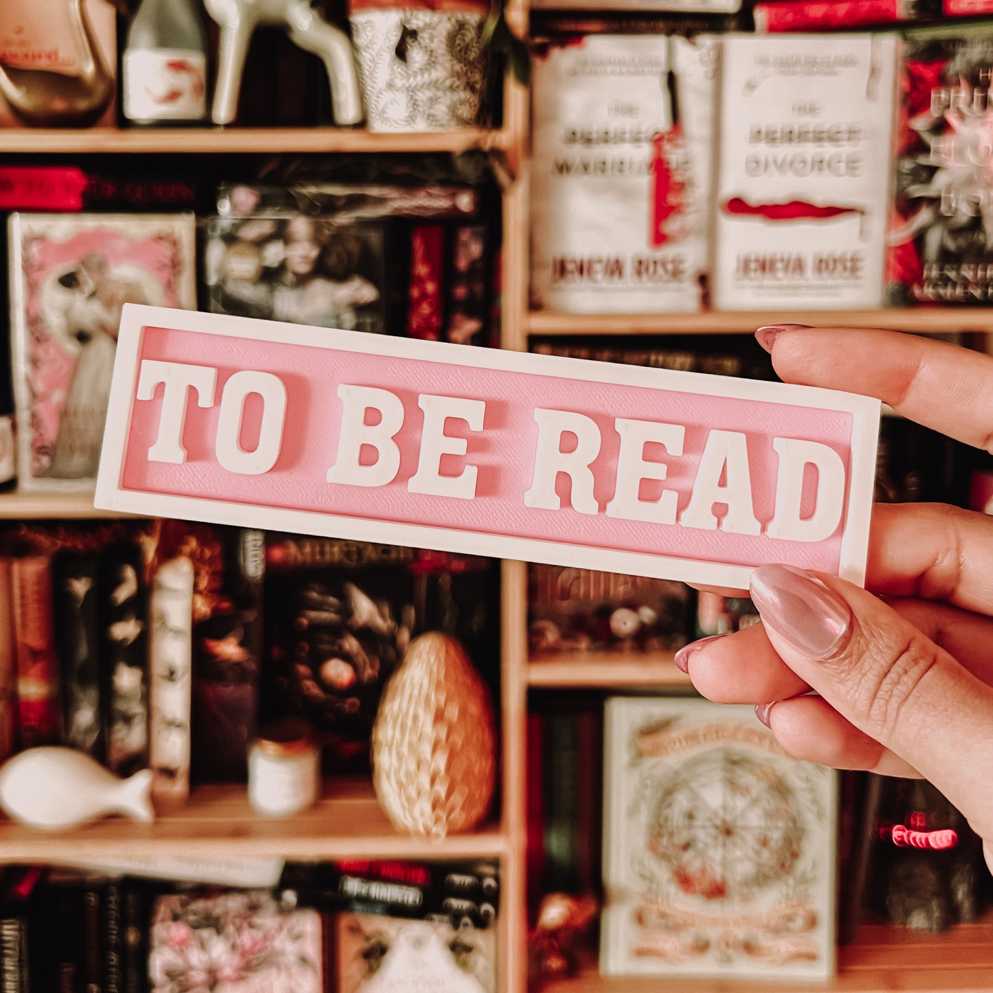 Person holding a 'TO BE READ' sign in front of a bookshelf filled with books and decorative items.