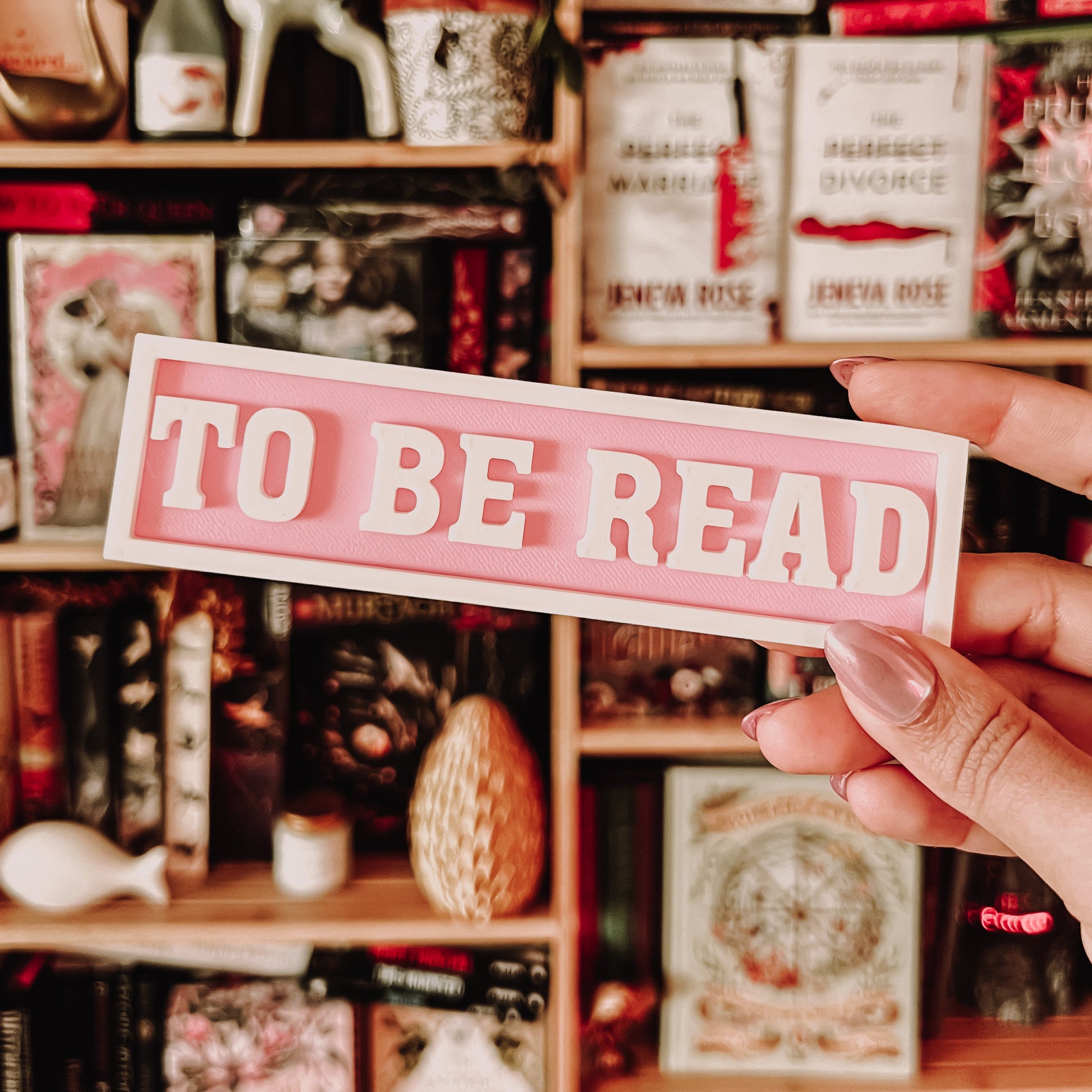 Person holding a 'TO BE READ' sign in front of a bookshelf filled with books and decorative items.