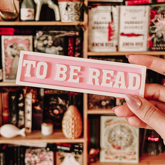 Person holding a 'TO BE READ' sign in front of a bookshelf filled with books and decorative items.