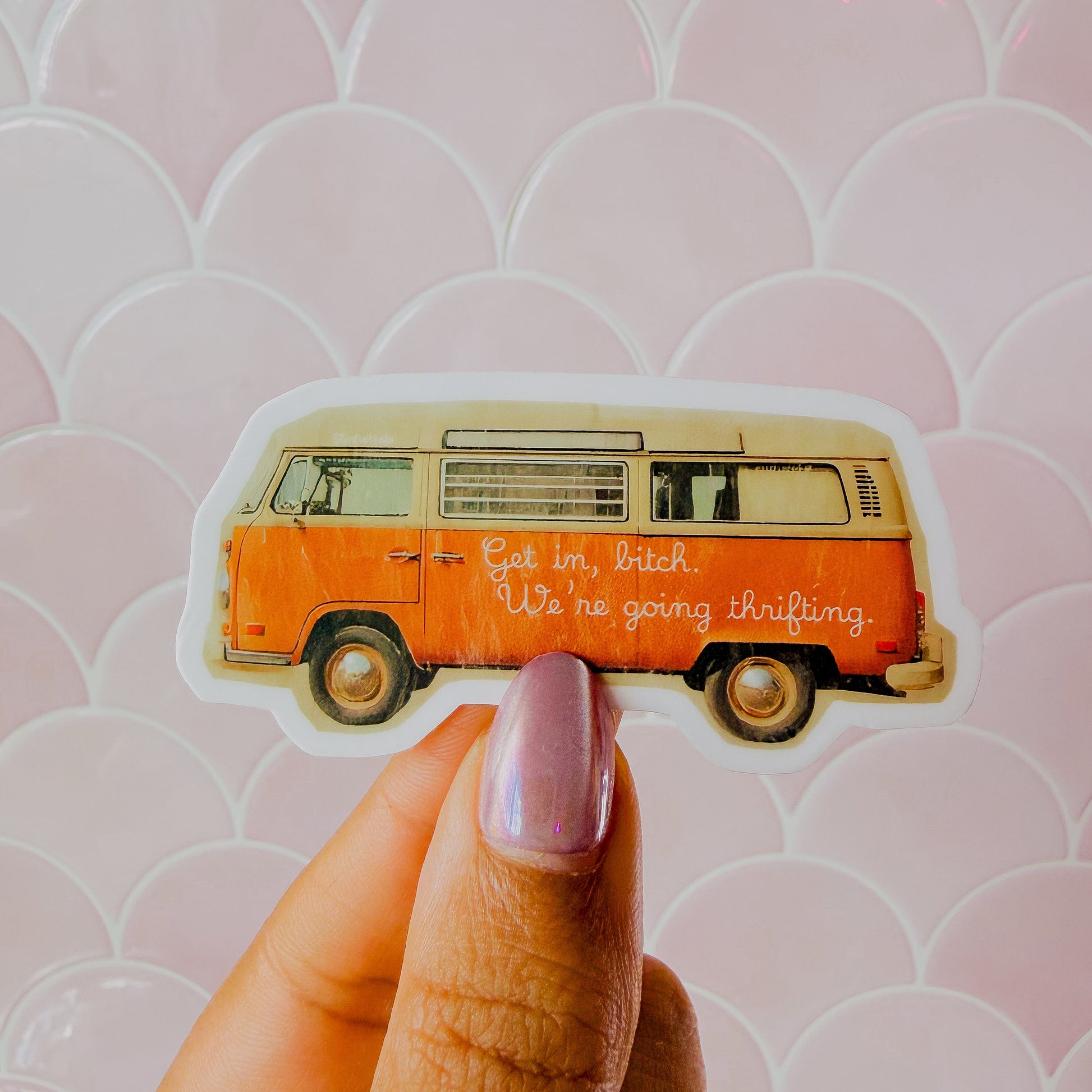 Hand holding a sticker of an orange van with text against a pink patterned background