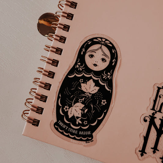 Pink notebook with a decorative sticker of a matryoshka doll on a light gray background